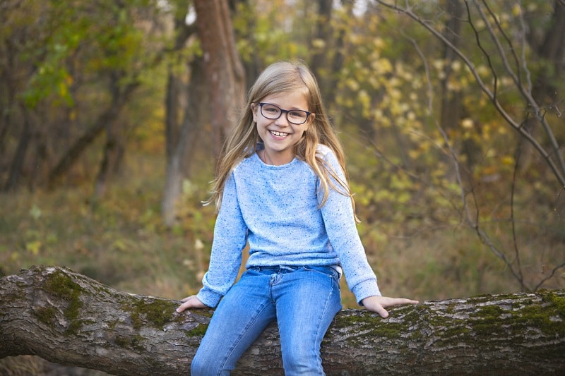 Cute young girl in jeans, t-shirt, and eyeglasses, sitting on a log in the woods and smiling